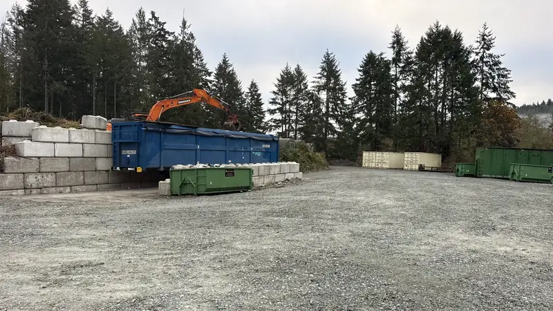 Orange excavator loading a blue bin in the Salt Spring Garbage drop-off yard, with green rental bins behind