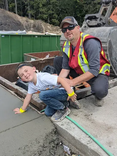 Owner Cory Kohlman crouching with his kid beside a fresh concrete pad at the Salt Spring Garbage yard
